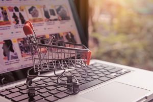 Criar site de vendas: Um pequeno carrinho de compras em um teclado de laptop, com uma tela de computador desfocada ao fundo exibindo um site de compras online.