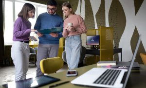Agência especializada em consultoria de marketing: Duas joven mulheres, uma vestindo um suéter lilás e jeans, e outra vestindo um suéter rosa e jeans, está ao lado de um homem que usa um suéter azul. Todos estão olhando para um tablet. Há um laptop em uma mesa em um ambiente de escritório decorado de forma colorida.