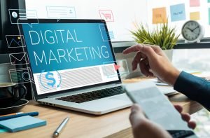 Serviços de marketing digital: Mãos de uma pessoa digitando em um laptop que exibe o texto "MARKETING DIGITAL" na tela, com vários ícones e elementos de marketing digital ao redor do laptop.
